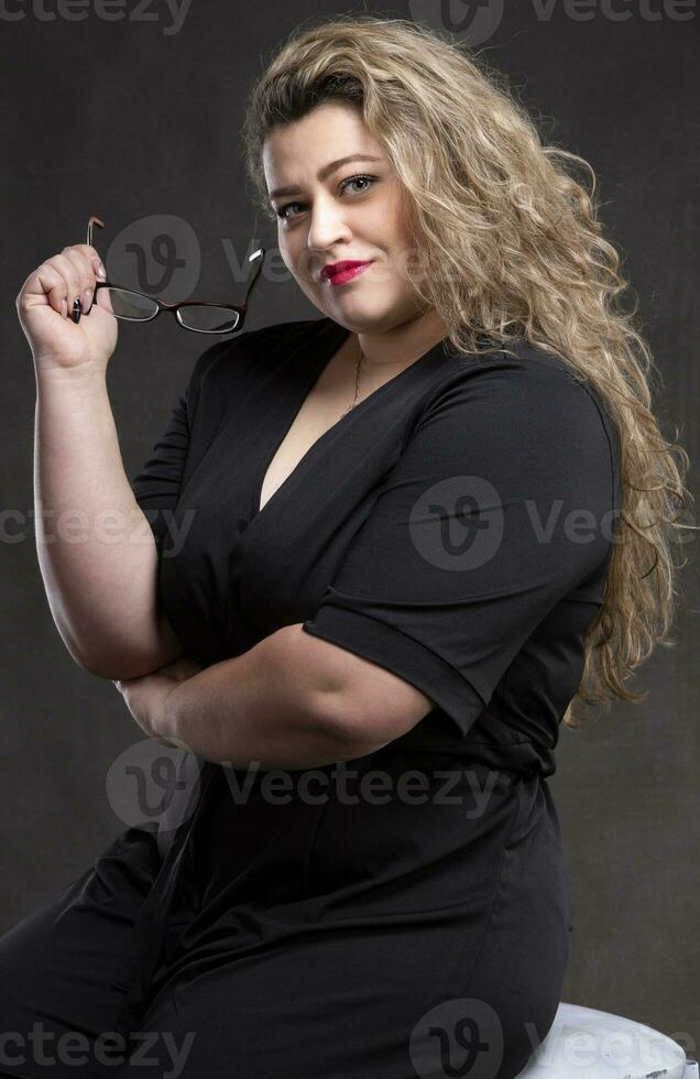 A beautiful fat woman with long curly hair holds glasses in her hands and looks at the camera and smiles. photo