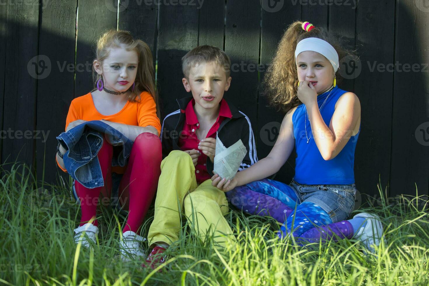 Beautiful model children in bright clothes sit on the grass near a wooden fence. photo