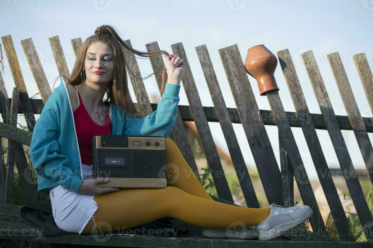 A beautiful country girl in bright clothes sits on a wooden bench with a cassette recorder. Woman in the style of the 90s. photo
