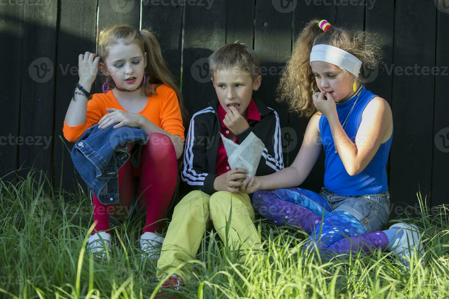 Cheerful kids, a boy and two cool girls sit on the grass near a wooden fence. photo