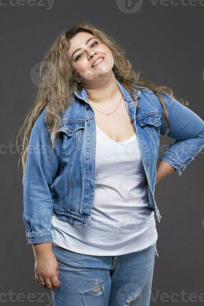 un hermosa grasa mujer en un mezclilla chaqueta mira a el cámara y sonrisas en un gris antecedentes. foto