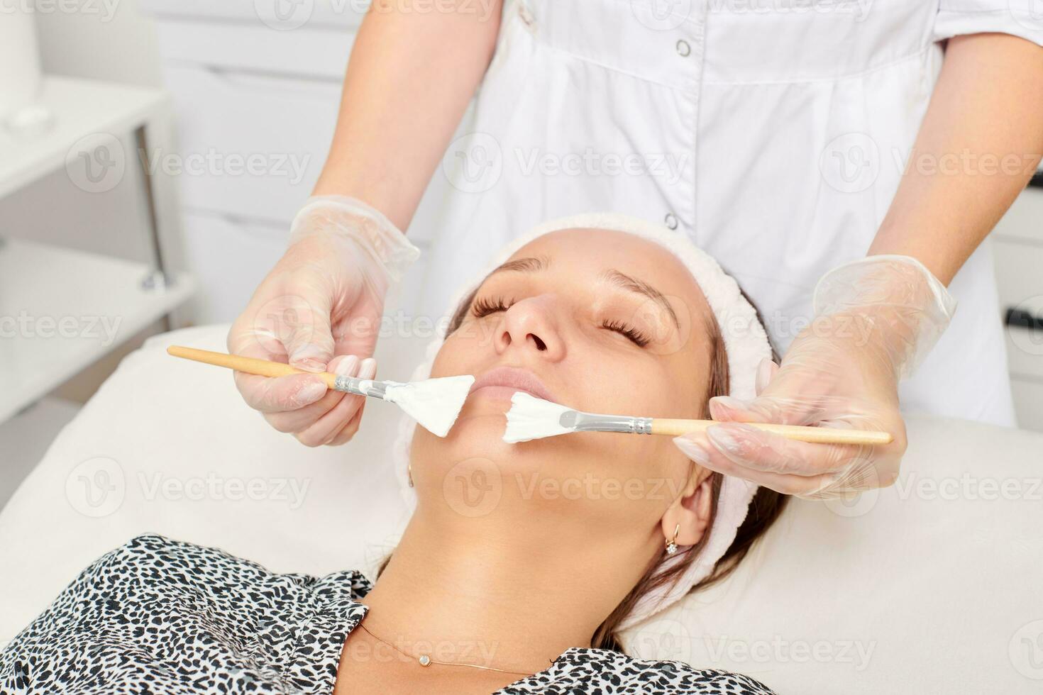 Beautician applying cosmetic cream mask on woman face for rejuvenation, procedure in beauty salon photo