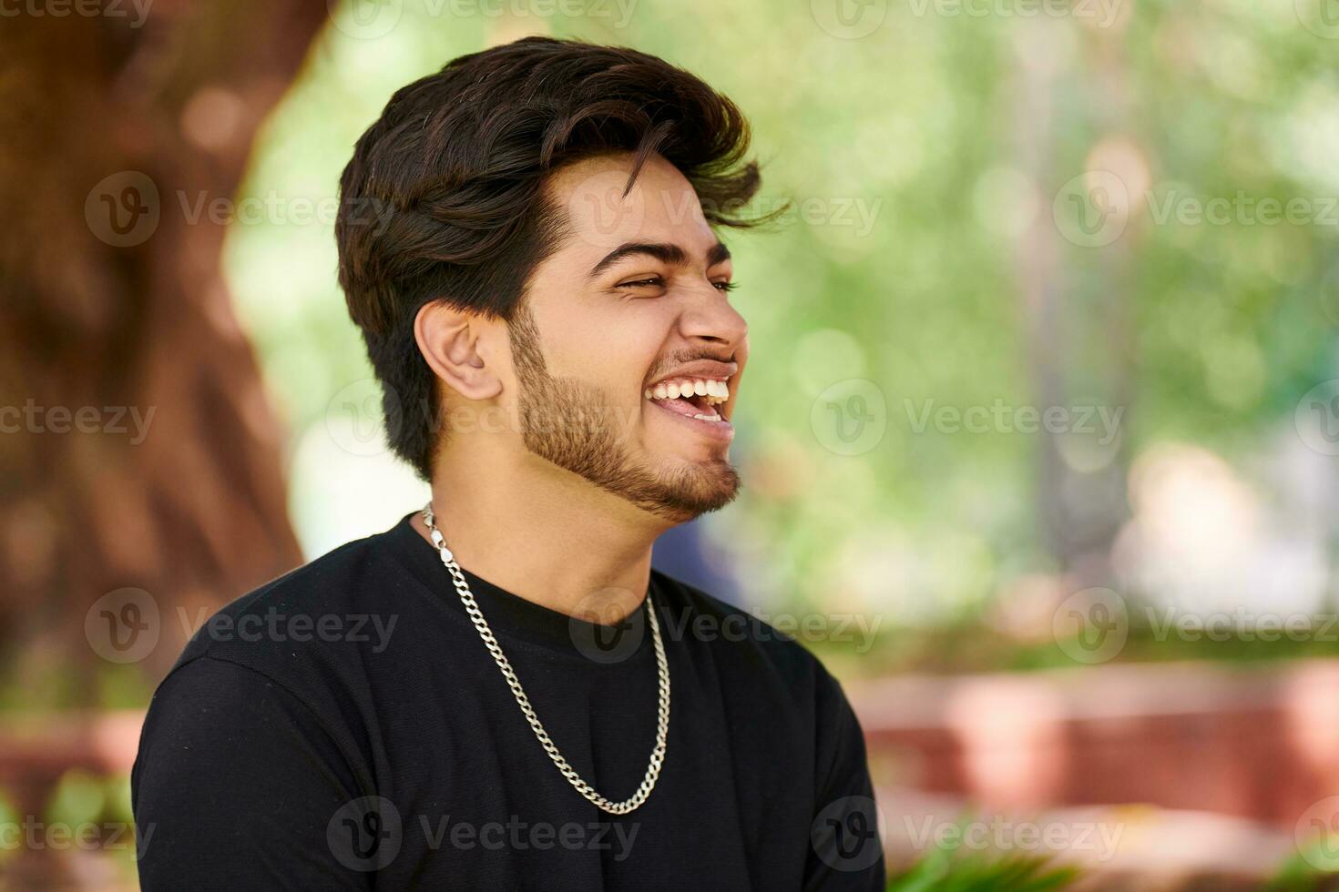 Smiling young indian man portrait in black t shirt and silver neck chain outdoor green public park photo