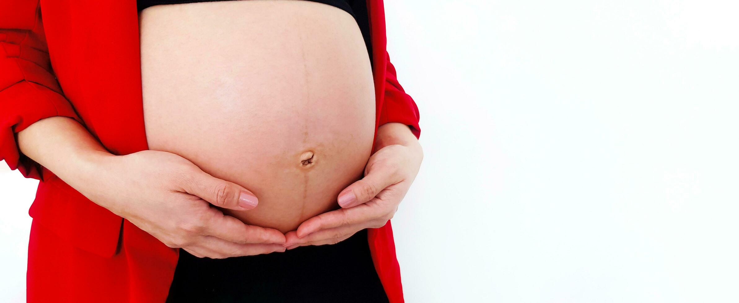 cerca arriba embarazada mujer vistiendo rojo traje sostiene manos en hinchado barriga aislado en blanco antecedentes y Copiar espacio con recorte camino. el embarazo 7-9 meses, maternidad, amar, expectativa y coche foto