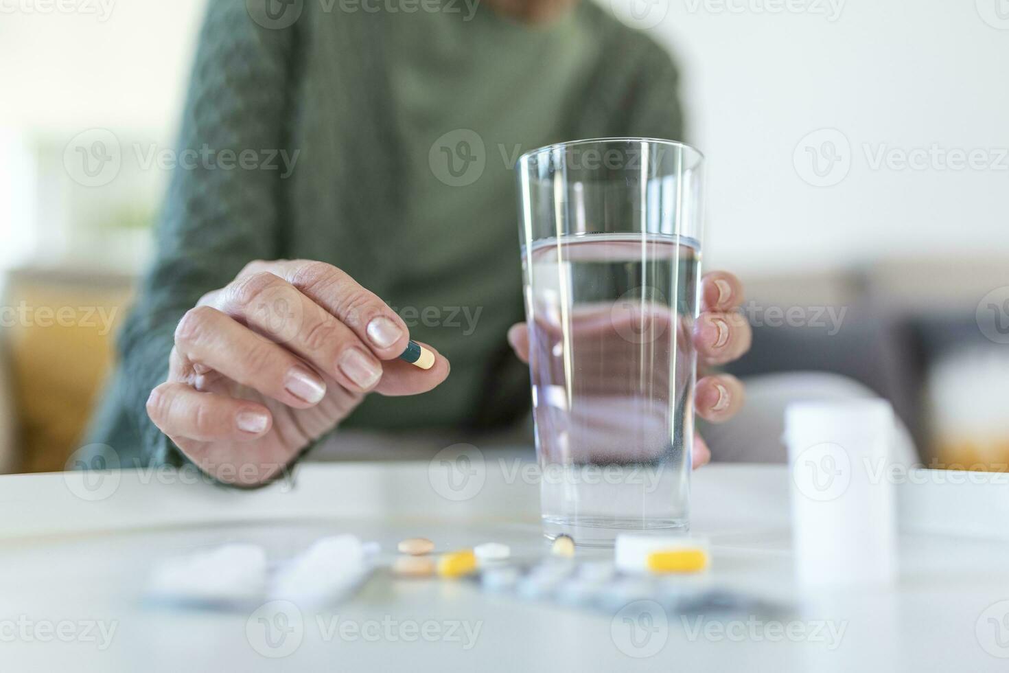 Healthcare concept. Cropped view of senior woman take vitamin pill, holding cup with water in hands, sitting alone behind wooden table photo