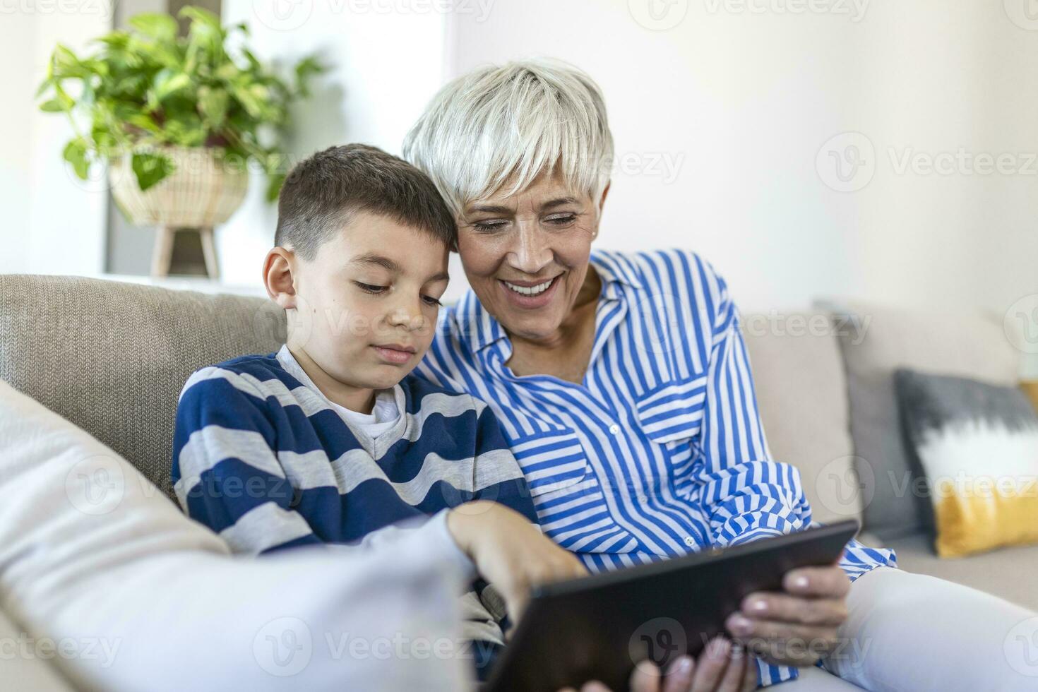 Happy grandmother and grandson using tablet together, sitting on cozy sofa at home, browsing tablet device apps, grandma with grandchild playing game, looking at screen, having fun photo