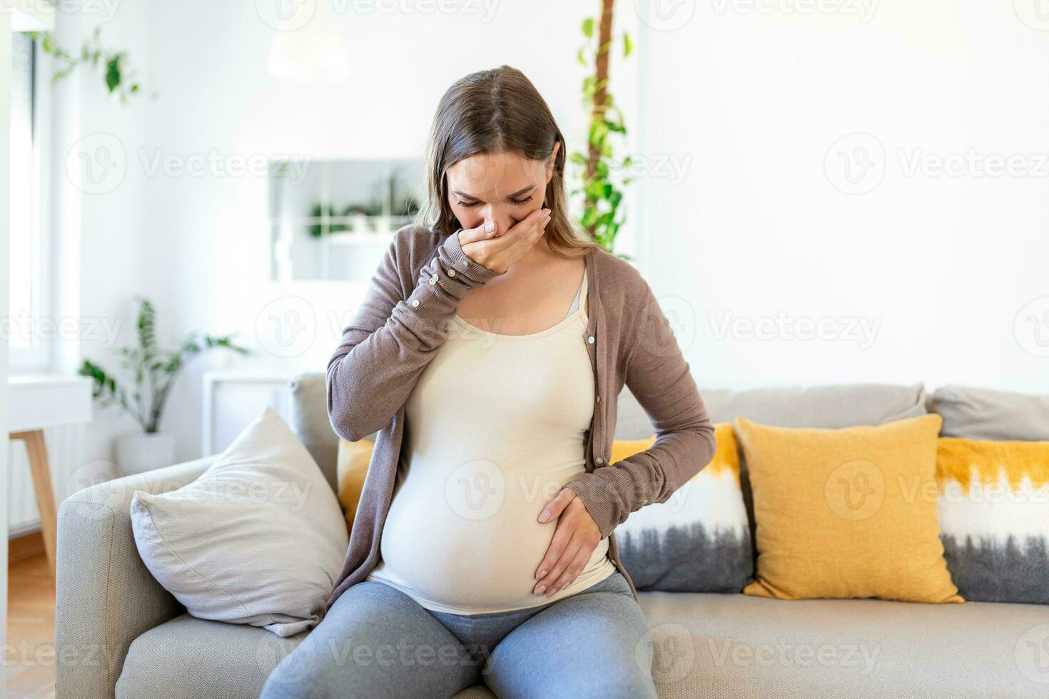 young pregnant woman suffering from toxicosis at home. maternity lady having morning sickness covering mouth with hands while sitting on couch in living room. mom expect unborn baby photo
