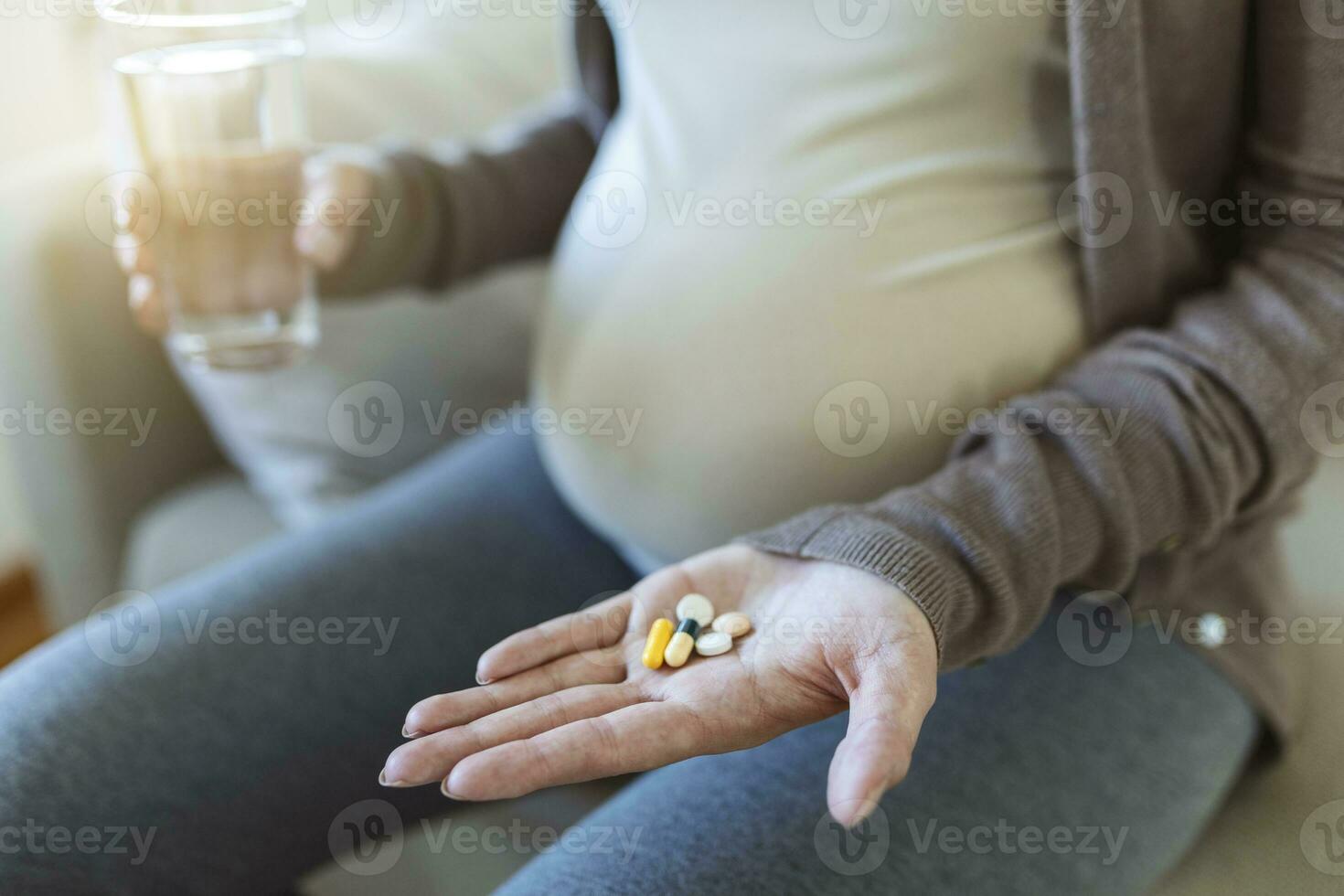 Healthcare concept. Cropped view of pregnant woman take vitamin pill, holding cup with water in hands, sitting alone behind wooden table photo