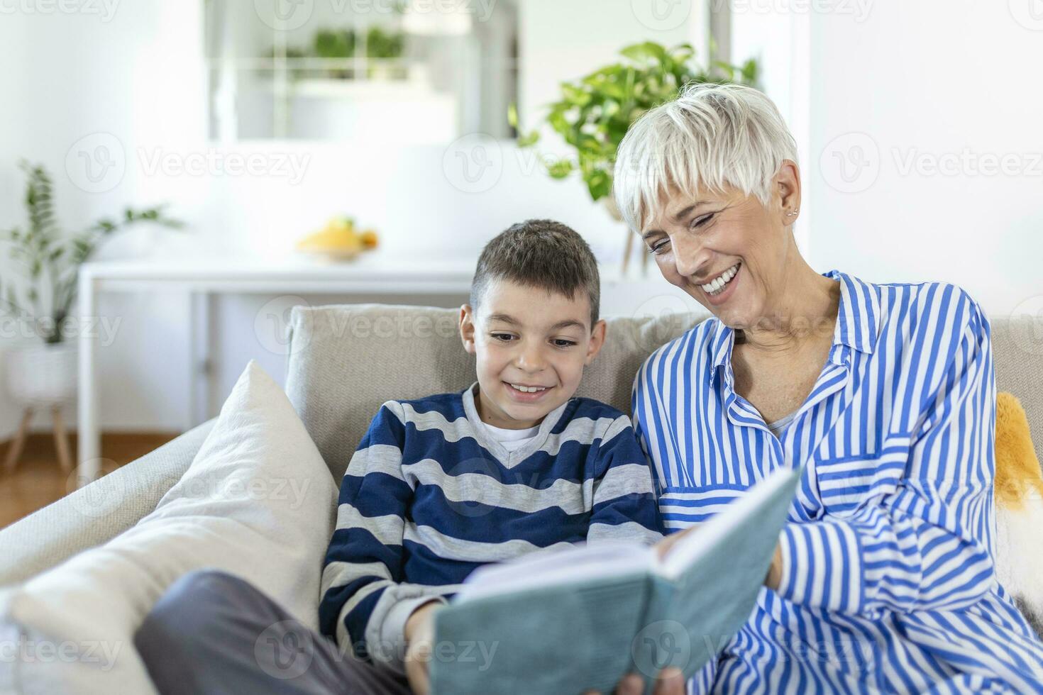 Cropped photo of happy gray-haired woman looking at book with her grandson indoors. They are smiling during their conversation. Visiting grandparents concept