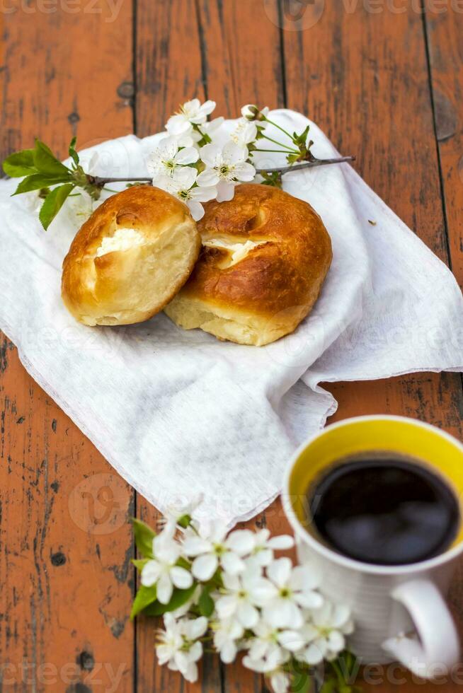 Baked open rolls and a cup of coffee on a dark, worn rustic wooden table. The composition is decorated with a twig with white flowers. Cherry tree flowers. Selective focus. photo
