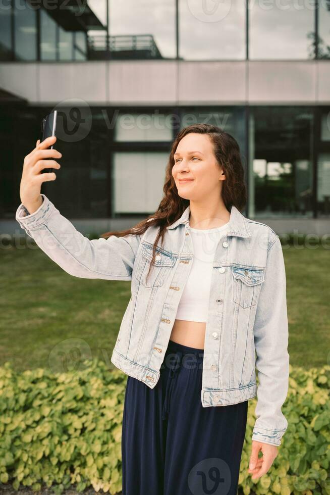 Young woman in white denim jacket taking selfie on street. Office building in the background photo