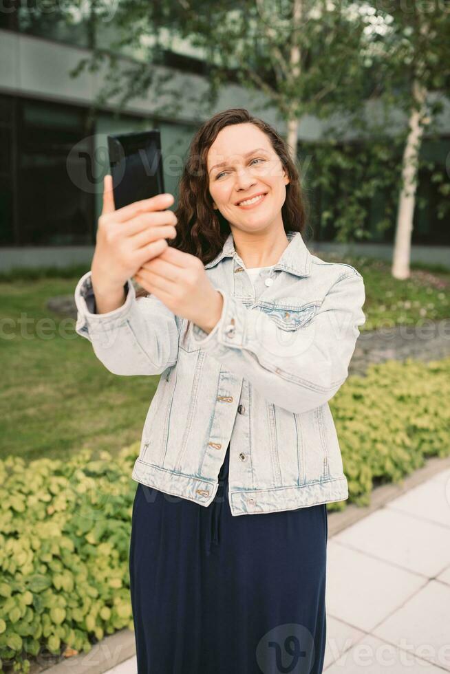Young woman in white denim jacket taking selfie on street. Office building in the background photo