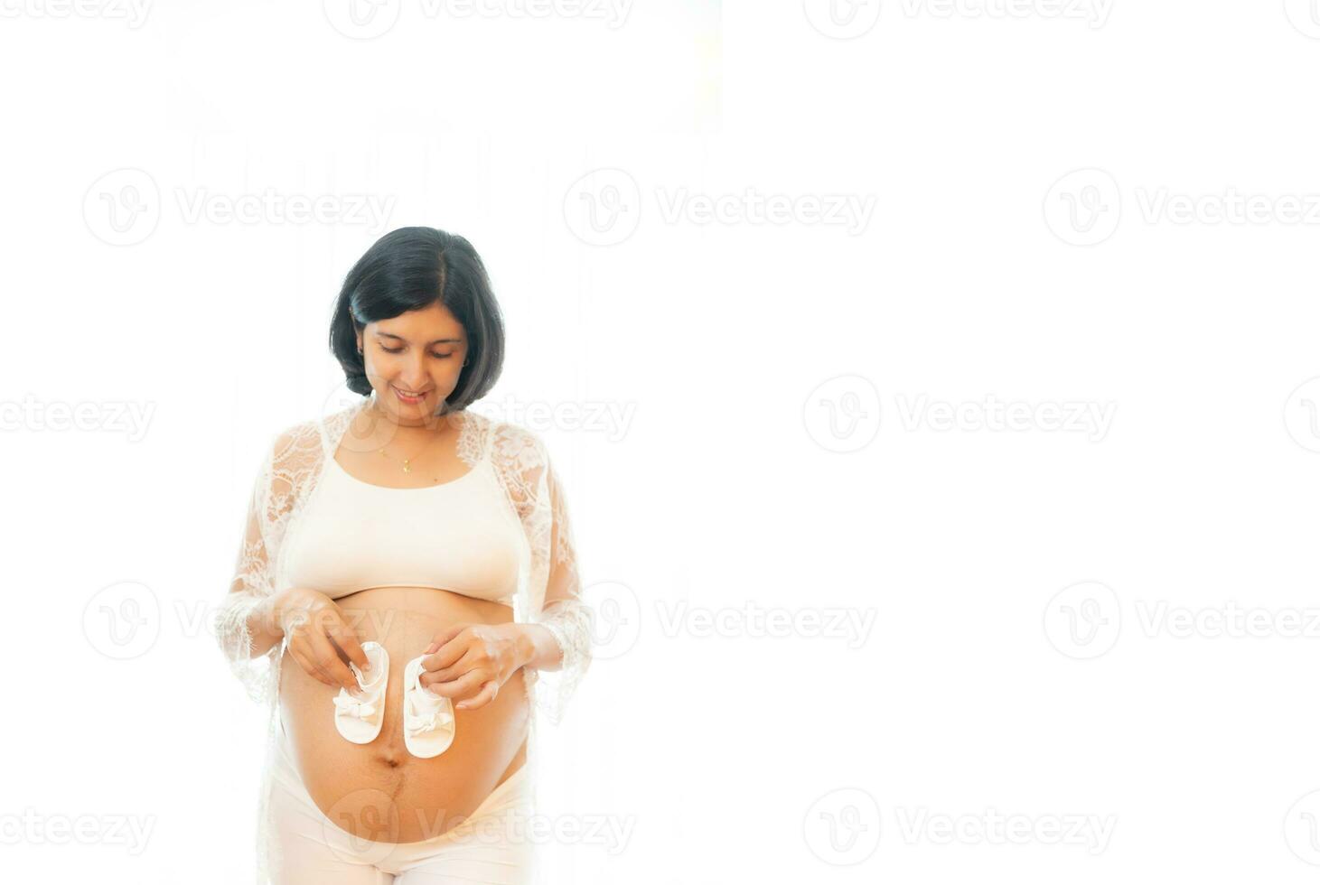 Closeup pregnant belly with cute tiny baby shoes. Concept of pregnancy, gynecology, preparation. Beautiful pregnant woman in shirt in white background. Woman holding big pregnant stomach. photo
