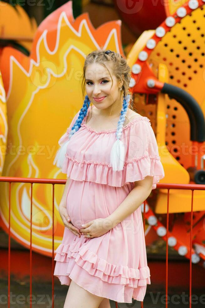 Cheerful pregnant young woman with blue pigtails and bright makeup walks in an amusement park photo