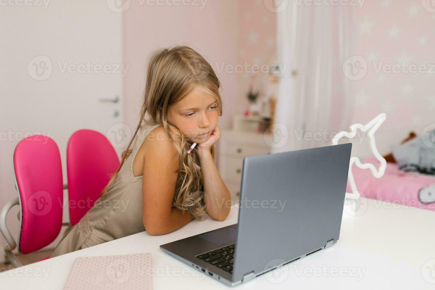 Thoughtful schoolgirl uses a laptop, looks at the monitor, studies online at home, sitting at the table. A child chatting with distance learning online. photo