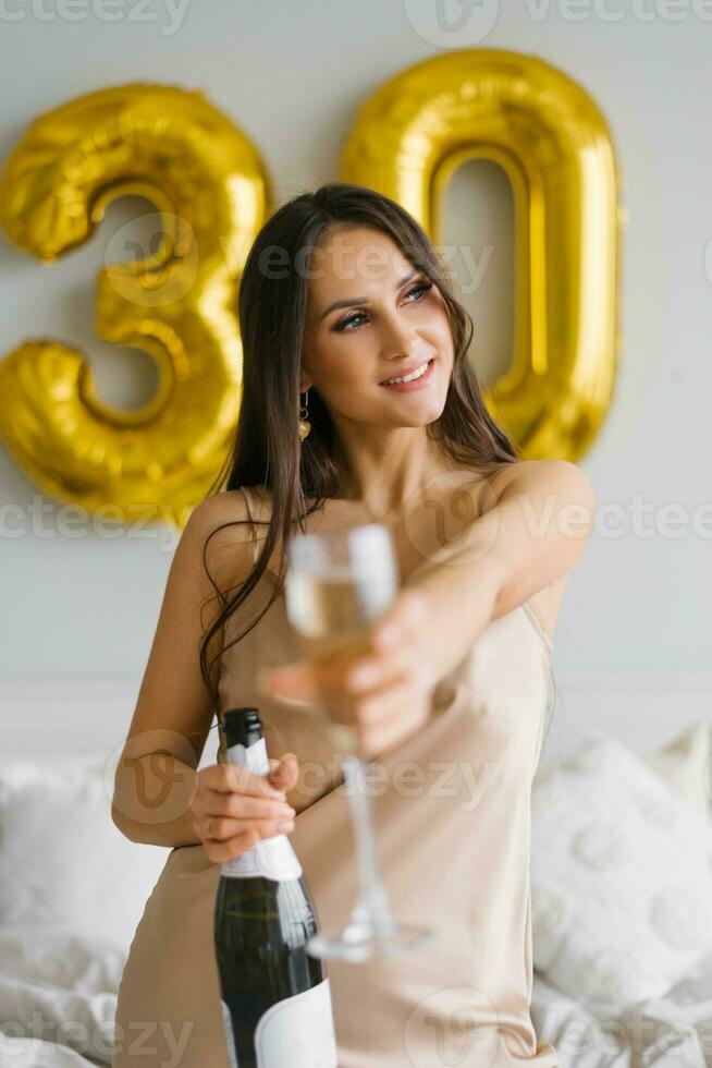 Pretty birthday woman celebrates thirty years and holds a bottle of champagne and a glass in her hands photo