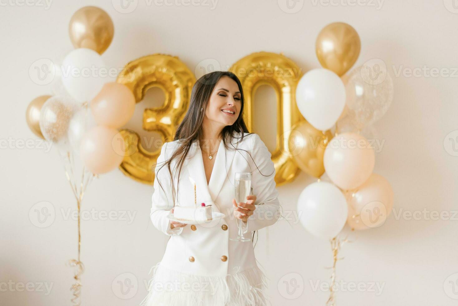 Positive woman celebrating anniversary dressed in stylish feather dress makes a wish with birthday candle cake around inflated balloons at home photo