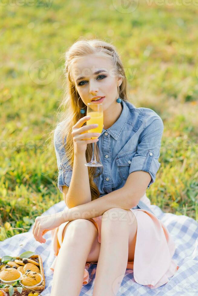 joven mujer en un azul mezclilla camisa y rosado falda en el jardín a un picnic participación un vaso de jugo en su mano foto