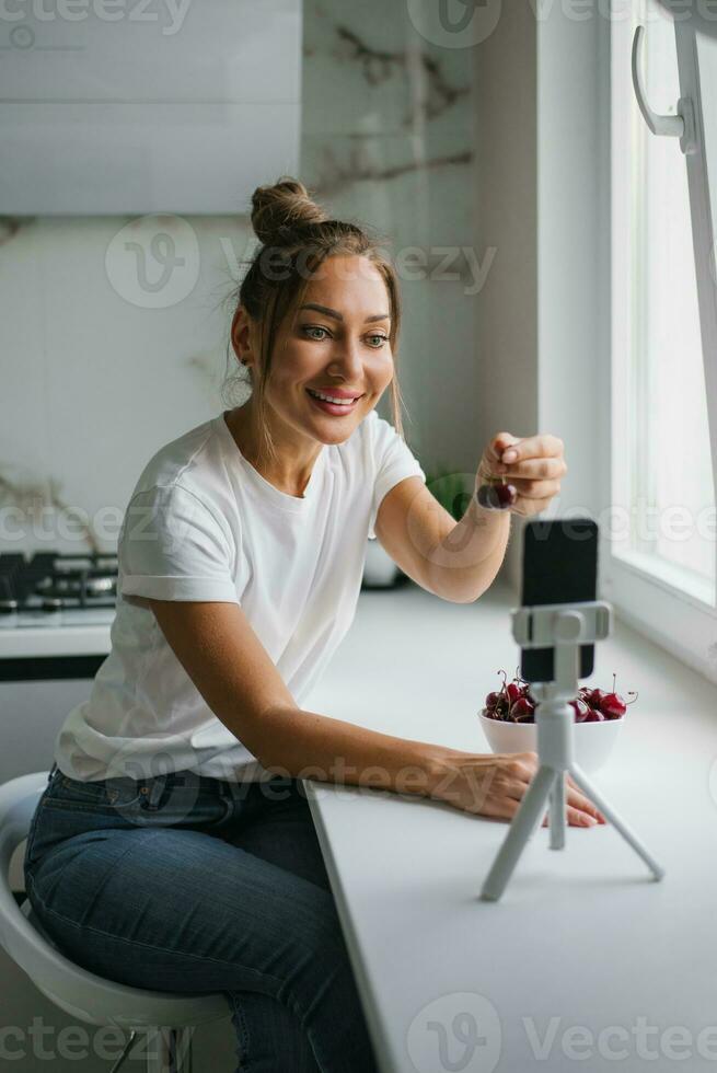 Beautiful young woman nutritionist conducts an online video call via smartphone with a friend to talk about the benefits of fresh cherries in the kitchen at home. photo