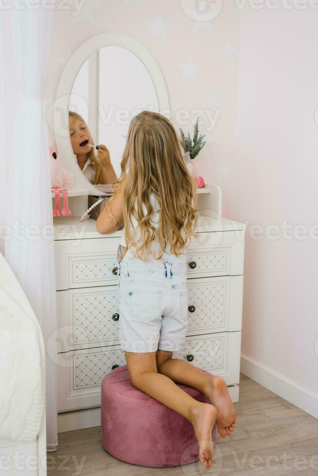 Pretty child girl paints her lips with glitter or balm in front of a mirror in her room photo