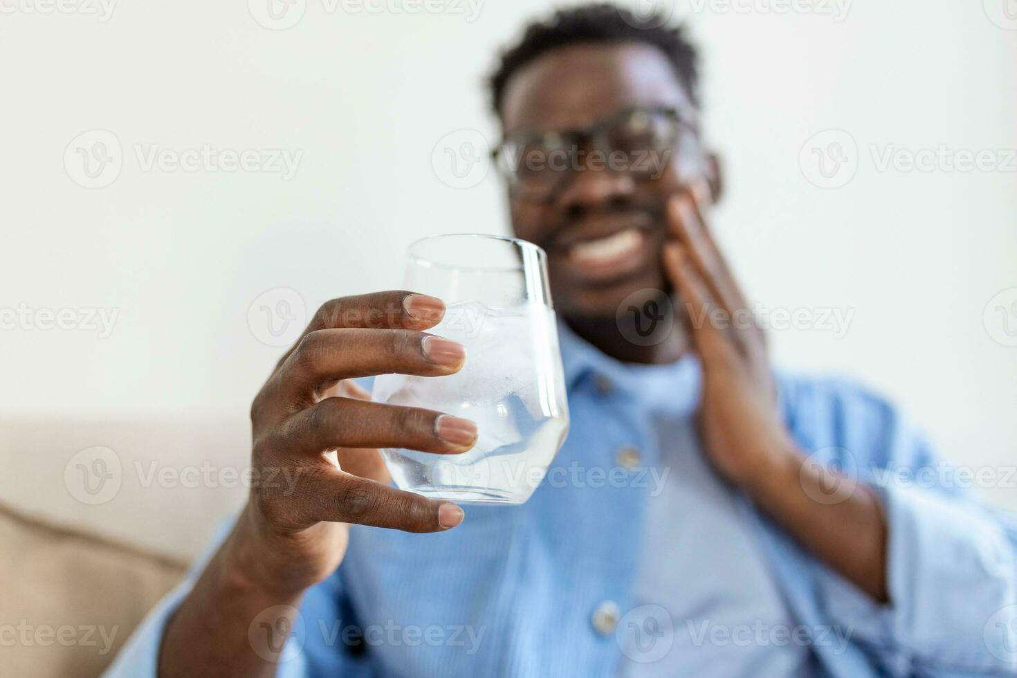 Young man with sensitive teeth and hand holding glass of cold water with ice. Healthcare concept. man drinking cold drink, glass full of ice cubes and feels toothache, pain photo