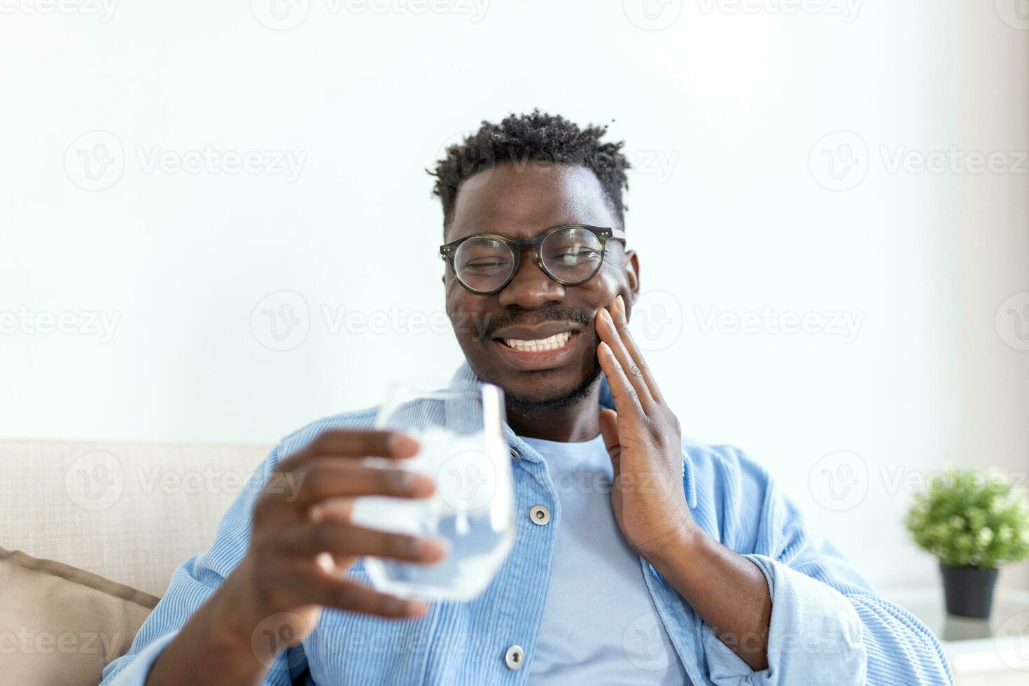 Young man with sensitive teeth and hand holding glass of cold water with ice. Healthcare concept. man drinking cold drink, glass full of ice cubes and feels toothache, pain photo