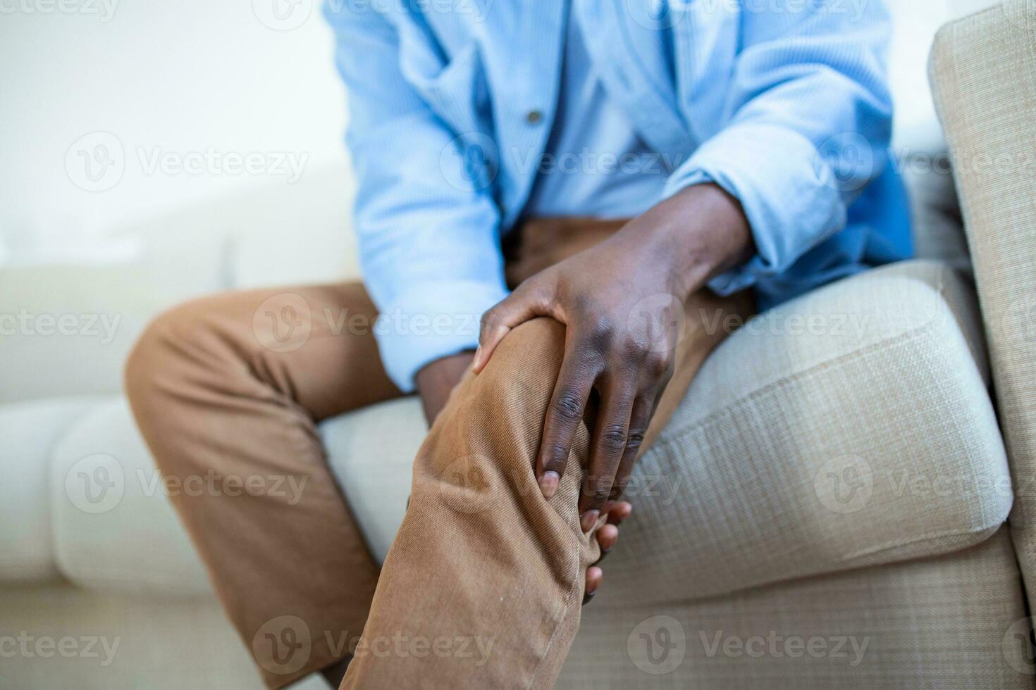 foto de africano hombre sentado en un sofá en el vivo habitación a hogar y conmovedor su rodilla por el dolor durante el día. hombre masajear su doloroso rodilla.