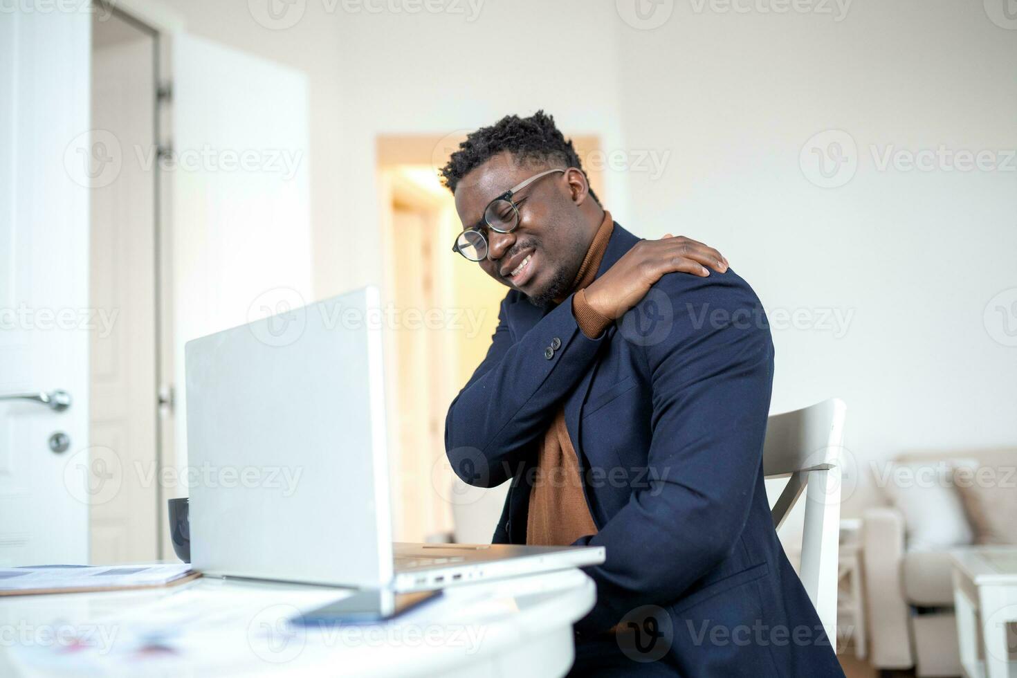 Tired African businessman working from home on his computer, feeling pain in his shoulder. man suffering from shoulder and back pain while sitting and working from home on laptop. photo