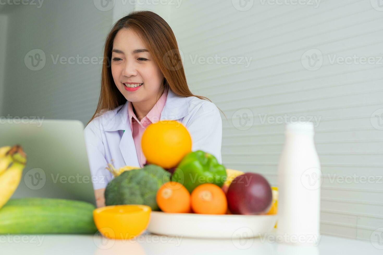 Portrait of Asian smiling female nutritionist typing on laptop computer for balanced corrective diet plan for patient or weight loss advice. Concept of healthy lifestyle, healthy food, dieting photo