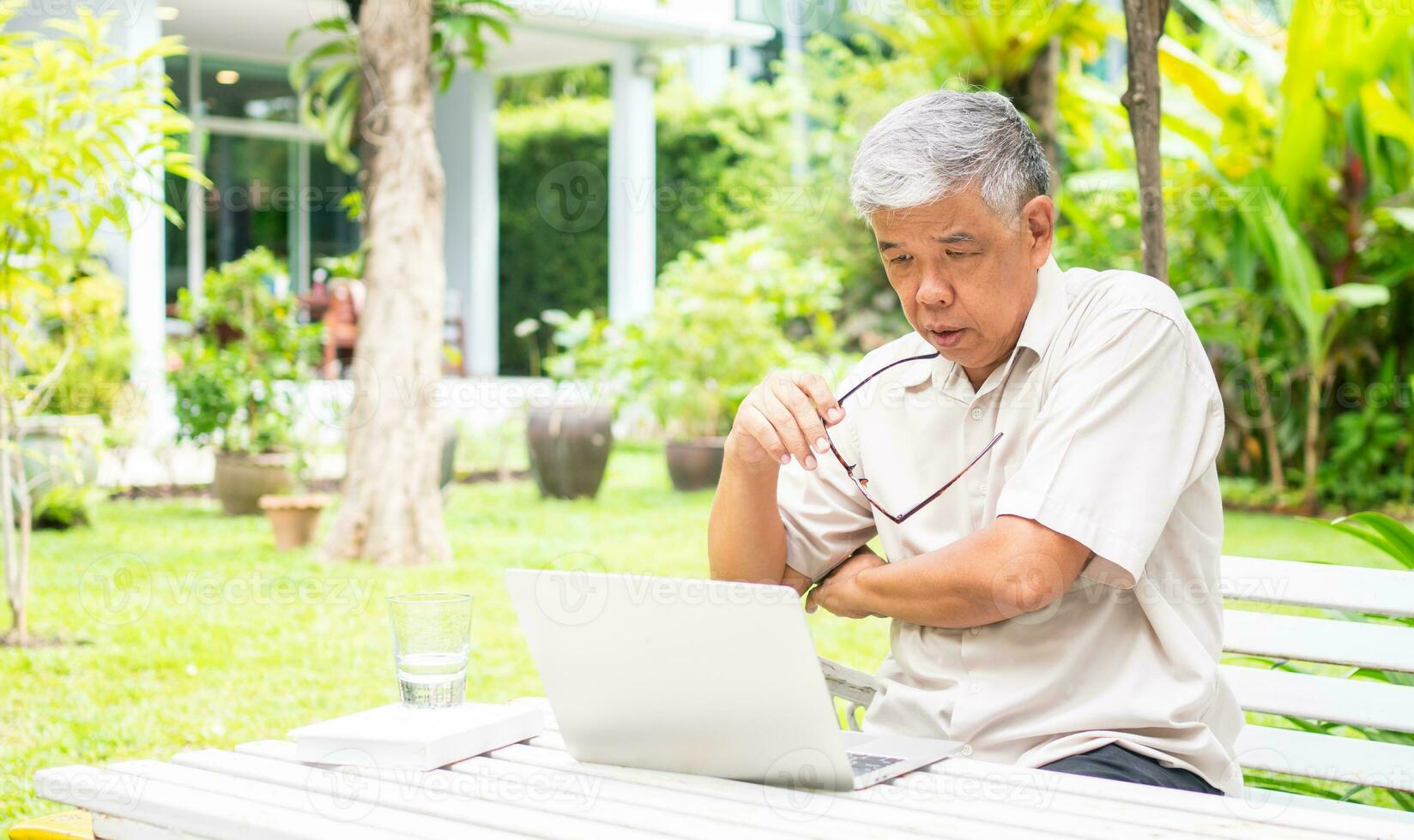 Portrait of old elderly Asian man using a computer laptop in the backyard for learning new skill after retired. Concept of no Ageism and not be late for learning. photo