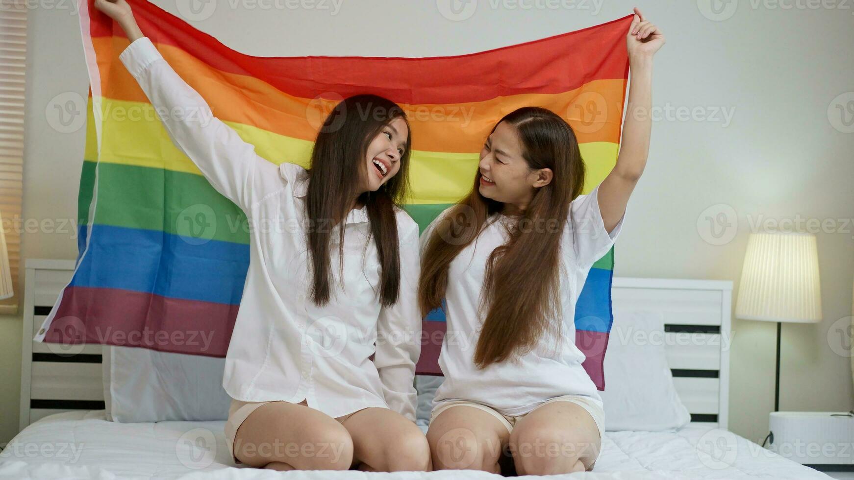 Portraits of happy Asian lesbian couple sitting with rainbow flag sitting bed beside the window. Cheerful two young adult women living together with relationship concept. LGBTQ Lifestyle Pride Month photo