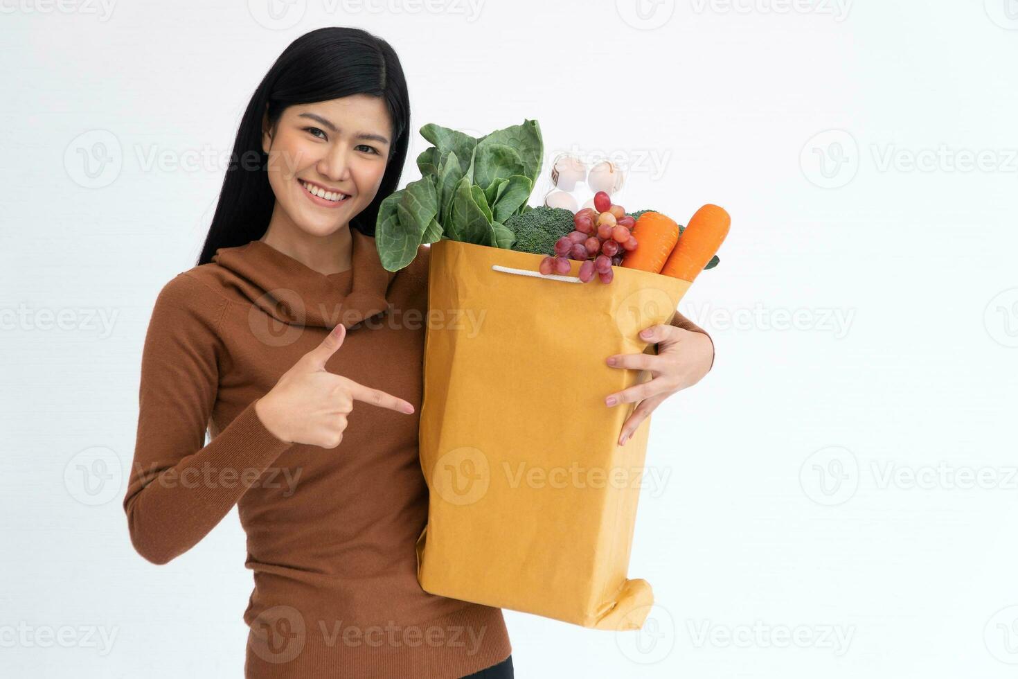 Happy Asian woman smiling and point the finger to a paper bag and carries a shopping bag after the courier from grocery came to deliver at home. Concept of Supermarket delivery for a new lifestyle. photo