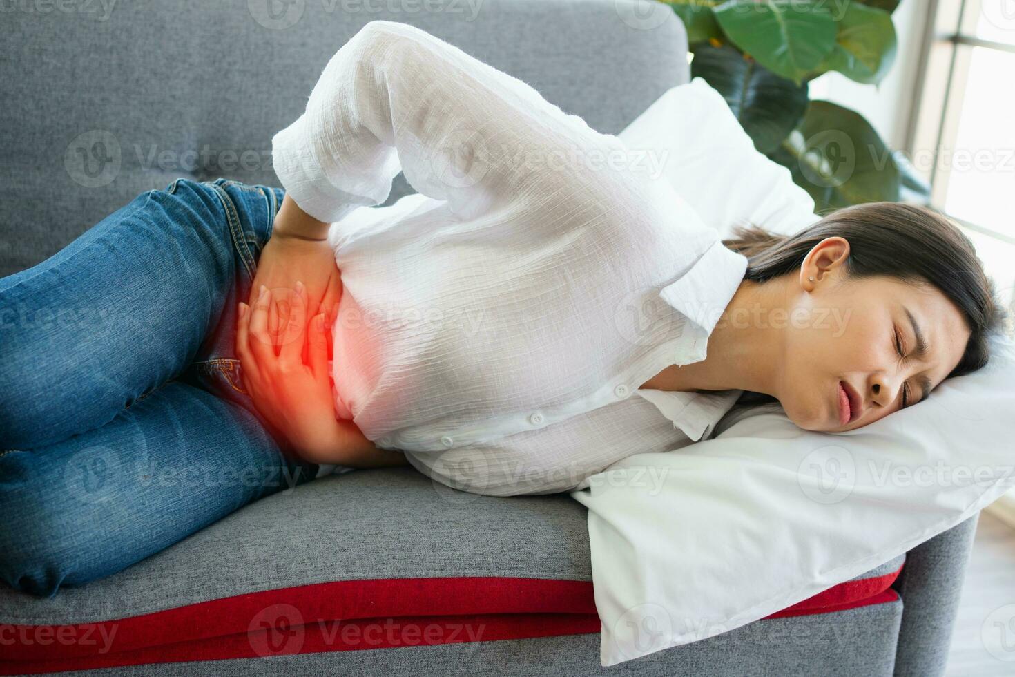 Unhappy Asian woman sitting on the sofa and holding on stomach suffering. Abdominal pain that comes from menstruation, diarrhea, or indigestion. Sickness and healthcare concept photo