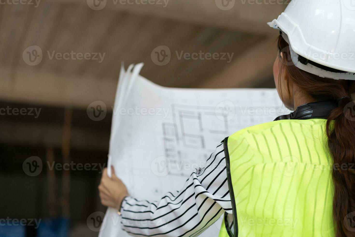 Asian engineer or Young woman Architect put on a helmet for safety and look at Blueprint for Inspect Building factory Construction Site on a construction site. Smart working woman concept. photo