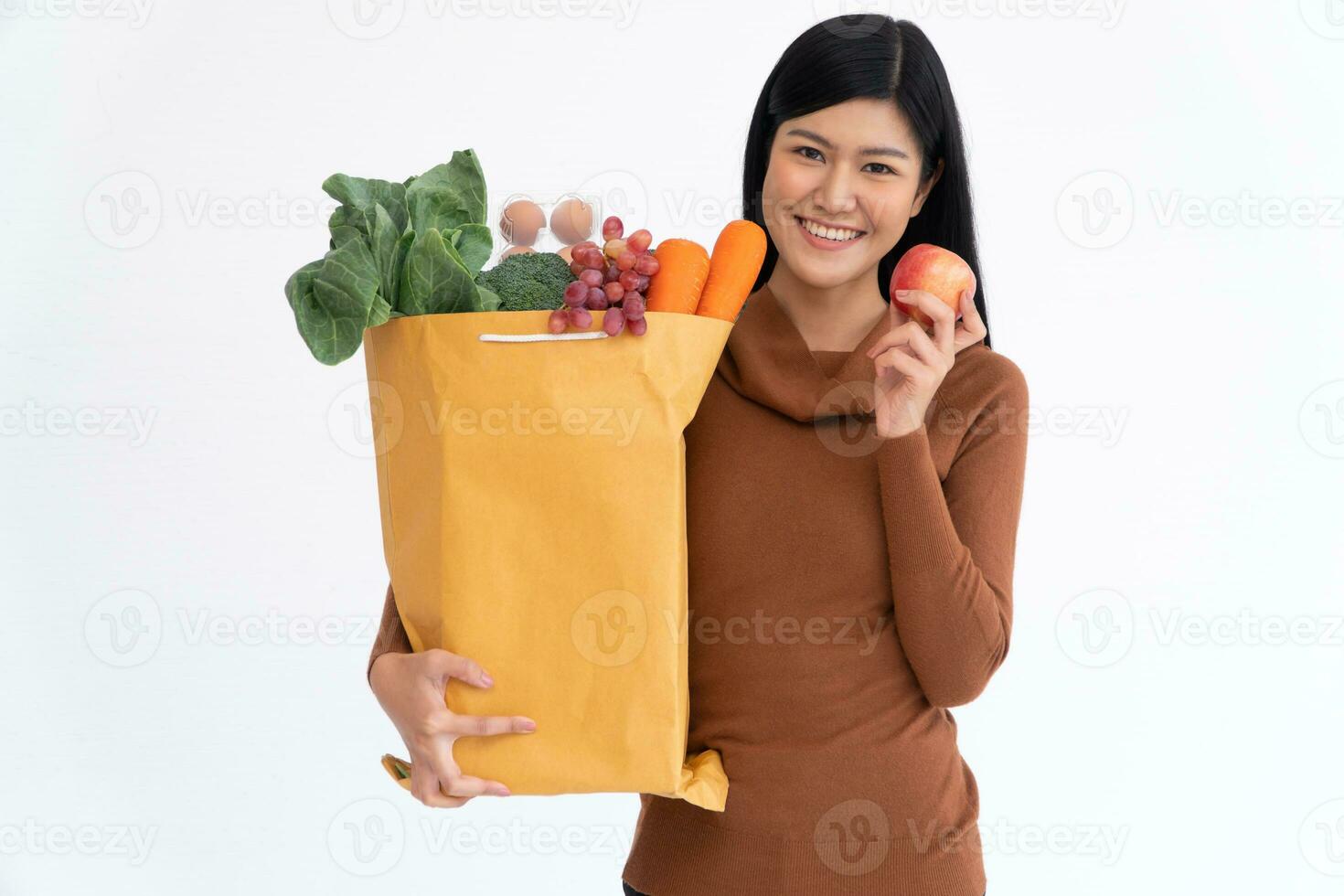 Happy Asian woman is smiling hold apple and carries a shopping paper bag after the courier from the grocery came to deliver his goods at home. Concept of Supermarket delivery for a new lifestyle photo