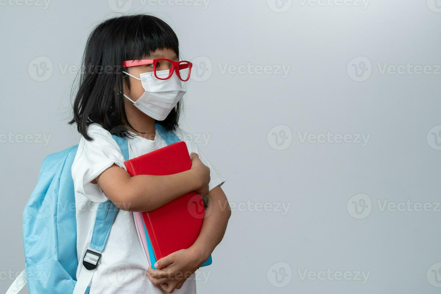 Happy Asian little preschool girl wearing red glasses holding books and red backpack on white isolated background. Concept of school kid and education in elementary and preschool, home school photo