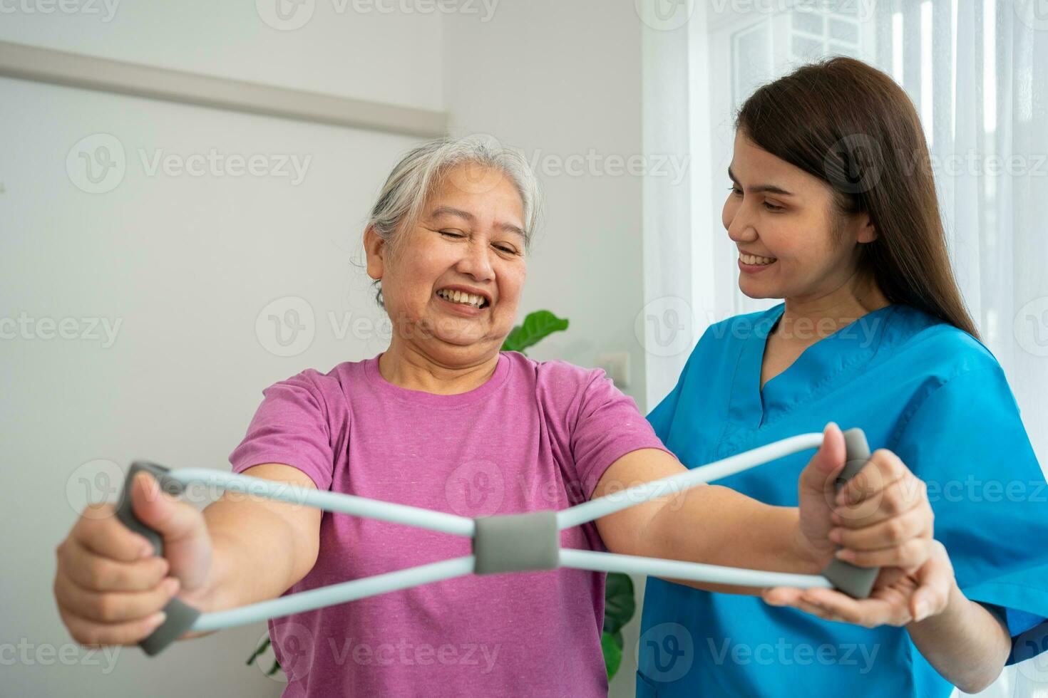 Happy elderly female exercising with rubber expander with Young physiotherapist for recovery arm muscles in a physiotherapy center. Concept of happy retirement with care from a caregiver and health. photo