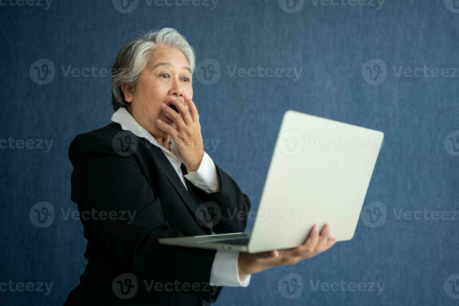 Portrait of intelligent senior businesswoman CEO manager holding laptop creating a presentation on isolated blue background. Old Woman is happy and smile after getting financial statements reports photo