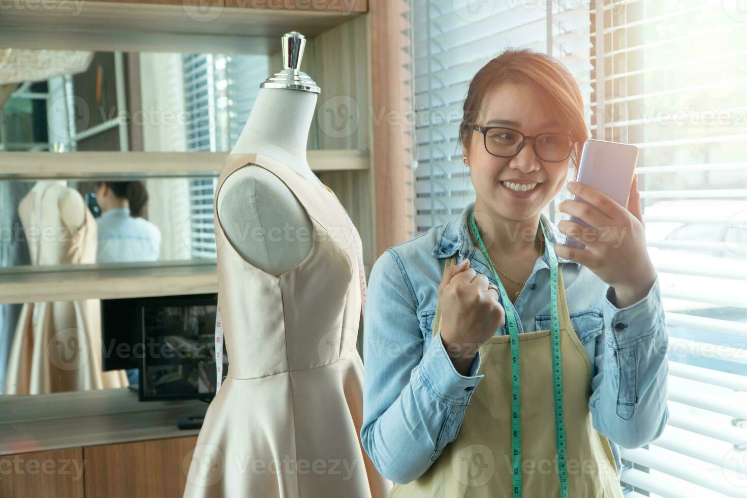 Happy young Asian woman dressmaker fashion designer uses a smartphone to accept orders from the customer in a showroom. Concept of success young entrepreneur in the fashion business. photo