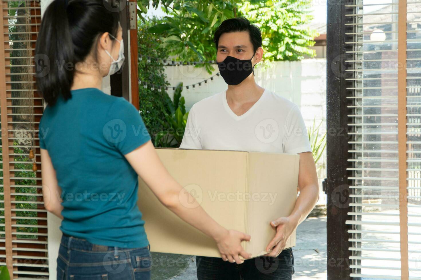 Asian man wearing a protective mask and delivering packages from grocery to young female customers at her home after online order. Concept of transportation service and express delivery and safety photo