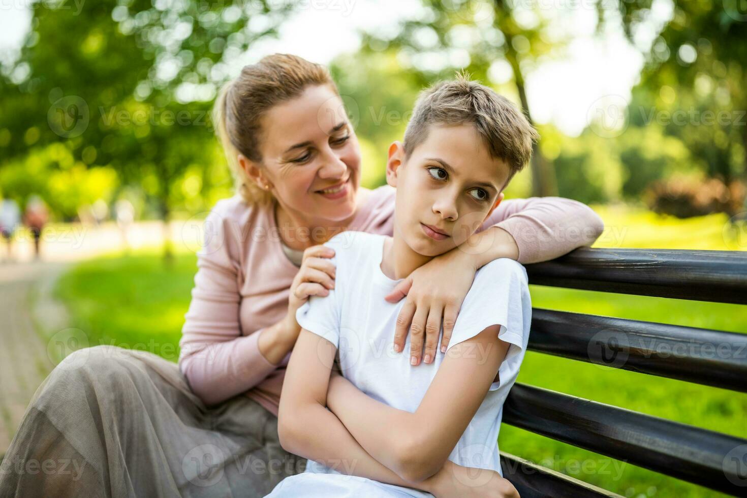 Happy mother is sitting with her son on bench in park. Boy is offended and pouting. photo