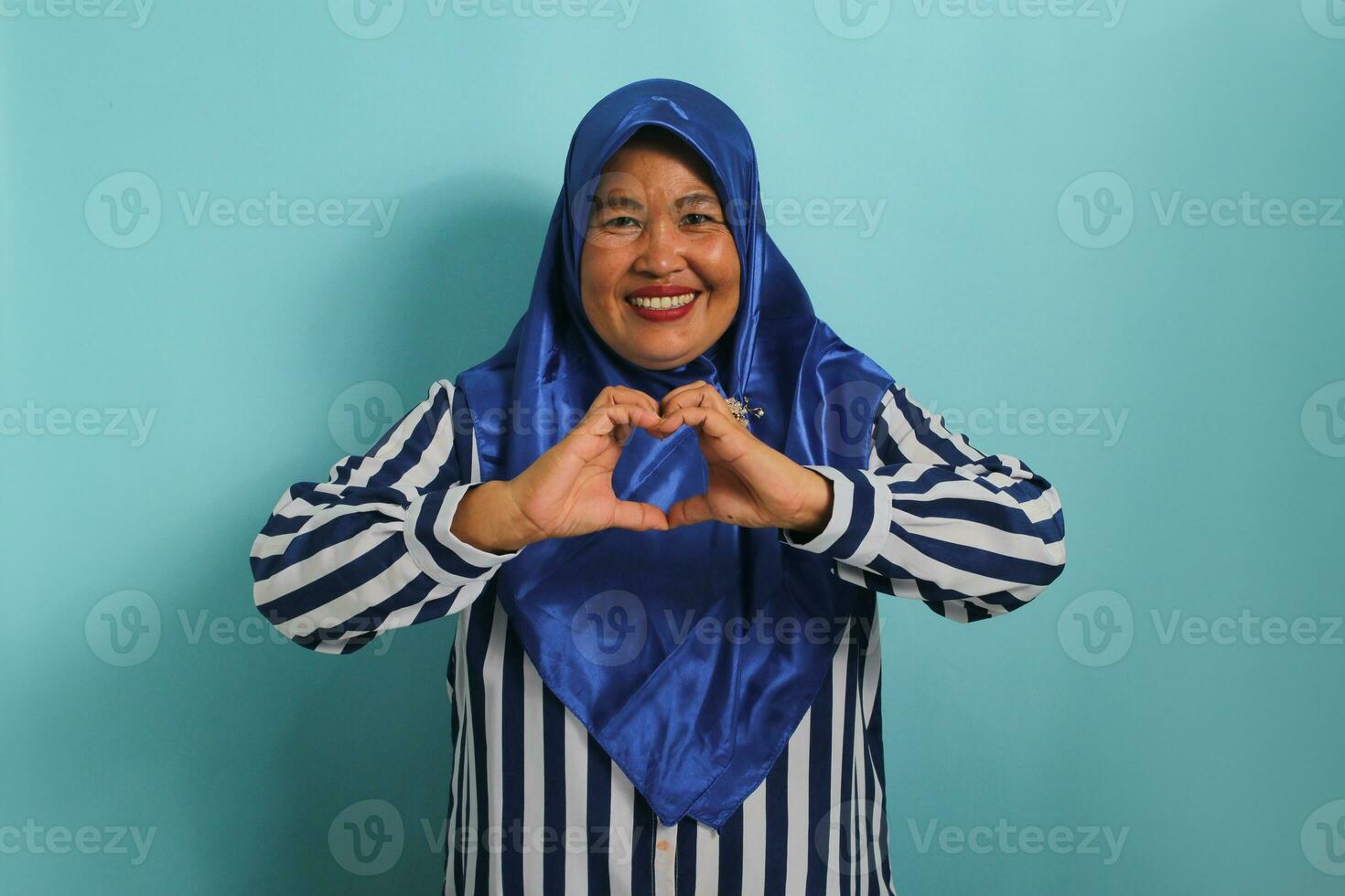 A smiling middle-aged Asian woman, in a blue hijab and striped shirt, makes a heart gesture with her hands, radiating happiness and care, isolated on blue background photo