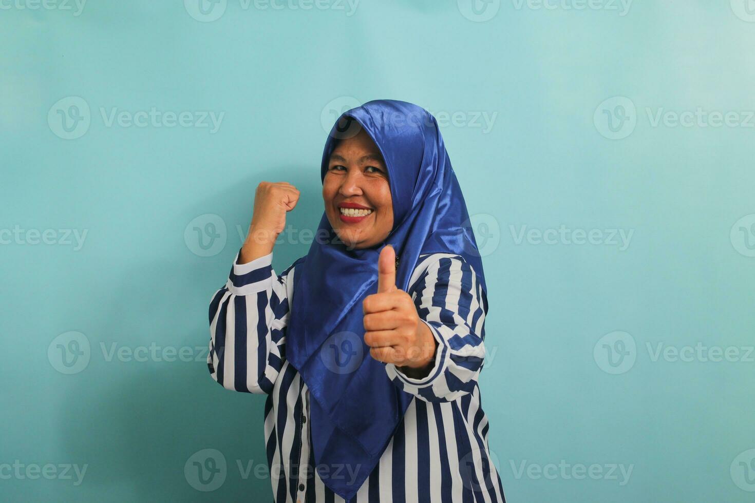 un emocionado de edad mediana asiático mujer en un azul hijab y a rayas camisa muestra un Pulgares hacia arriba y un bueno firmar, simbolizando éxito y expresando aprobación, mientras en pie en contra un azul antecedentes. foto