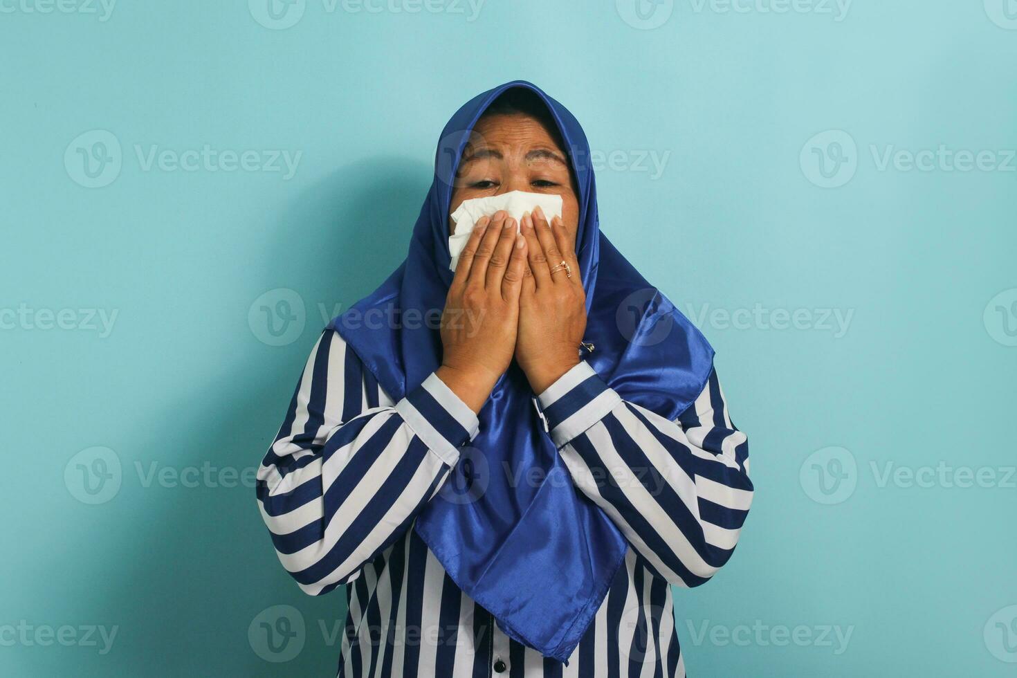 unhealthy middle-aged Asian woman, wearing a blue hijab and a striped shirt, is seen blowing her runny nose with a tissue while sneezing due to the flu. Healthcare and medical concept photo