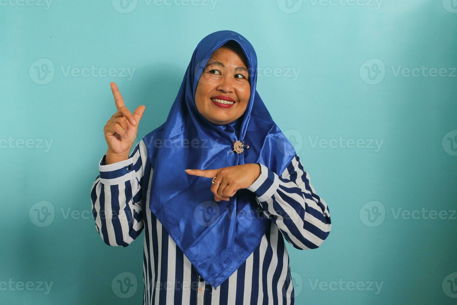 un entusiasta de edad mediana asiático mujer en un azul hijab y a rayas camisa puntos izquierda hacia un espacio de copia, aislado en azul antecedentes. foto