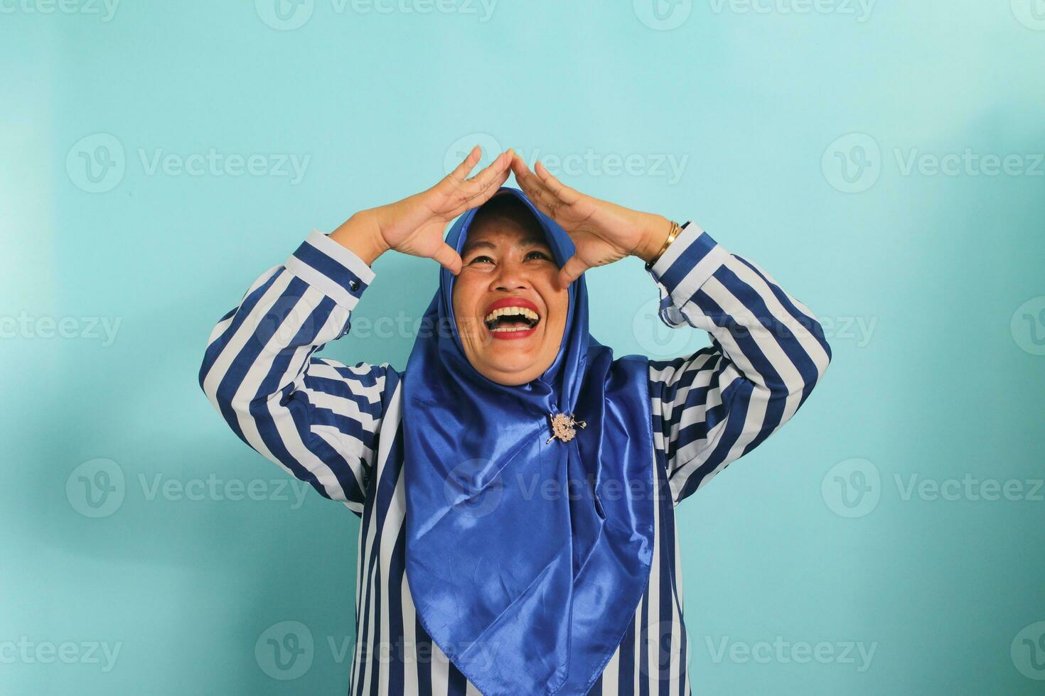 An excited middle-aged Asian woman in a blue hijab and striped shirt is making a home or roof gesture by holding her hands above her head while standing against a blue background photo