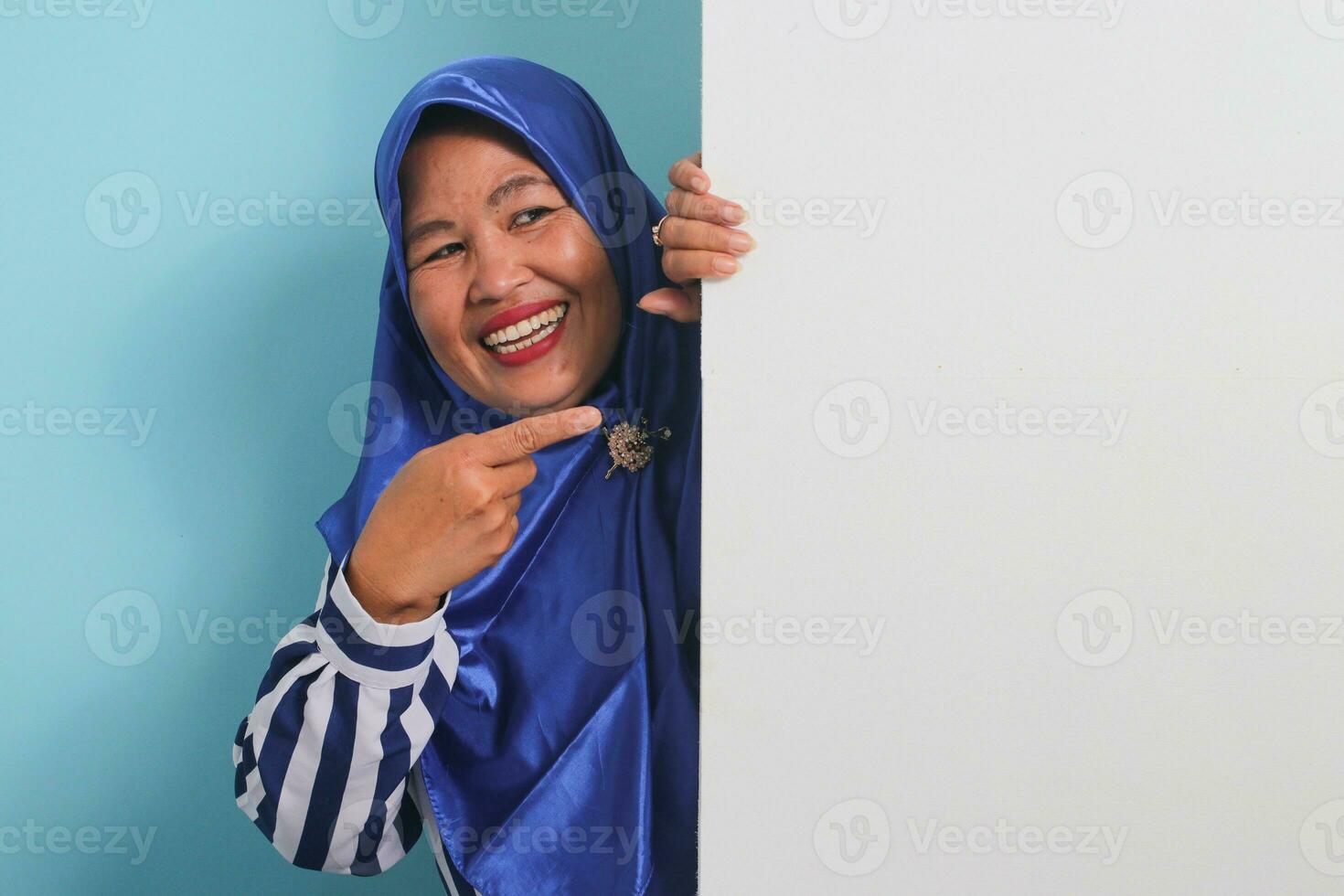 Excited middle-aged Asian woman in a blue hijab and striped shirt holds and pointing a blank white banner signboard with copyspace, offering ample space for your message, against a blue background. photo