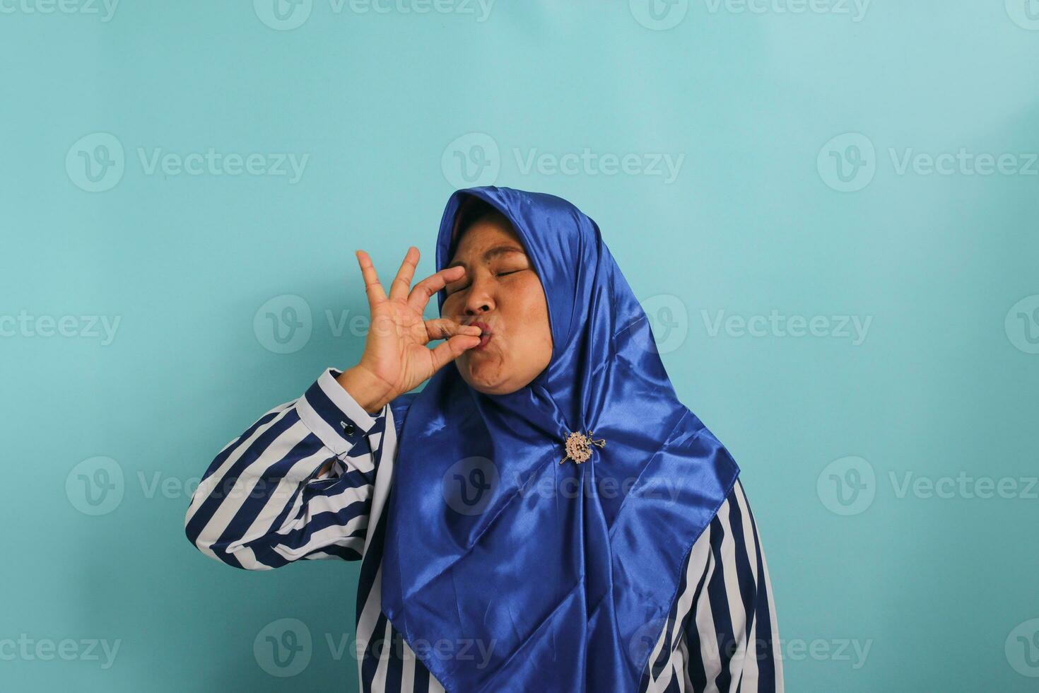 A happy middle-aged Asian woman, wearing a blue hijab and a striped shirt, is showing a delicious gesture by kissing her fingers and closing her eyes while standing against a blue background photo