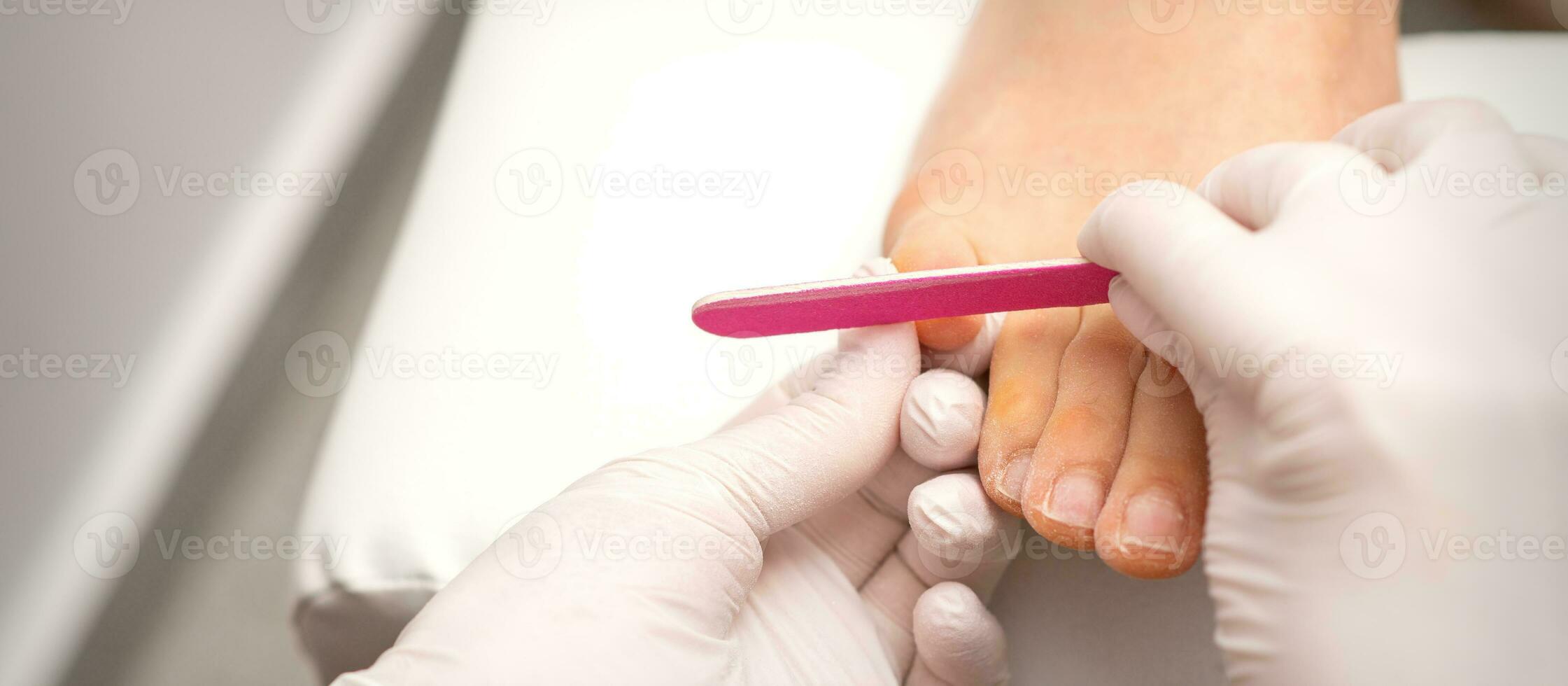 Pedicurist hands in protective rubber gloves filing toenails on feet with a nail file in a beauty salon. photo