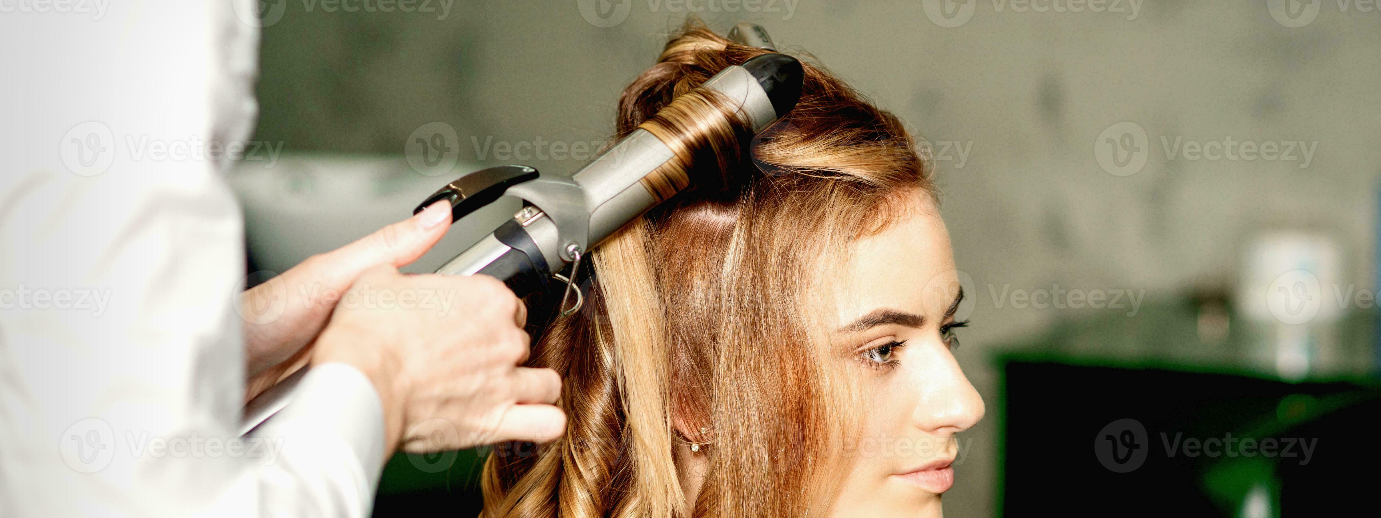 Hairdresser using curling tongs curls long brown hair on the young